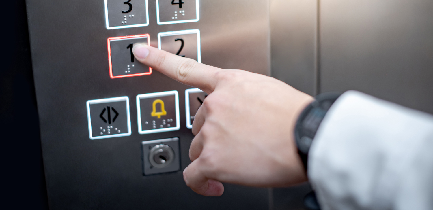 Lähikuva henkilöstä painamassa braille-merkein varustettua hissin painiketta
